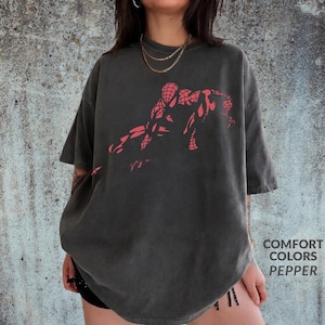 May include: A charcoal grey short-sleeve t-shirt featuring a red graphic print of a crouching superhero figure on the chest. The shirt has a crew neckline and a relaxed fit. The text COMFORT COLORS PEPPER is printed in the bottom right corner.