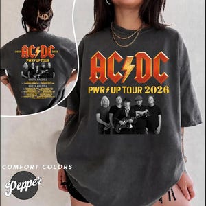 May include: A charcoal grey t-shirt featuring a large AC/DC PWR UP TOUR 2026 graphic on the front and back. The design includes the band name in bold orange letters, a lightning bolt, and a photograph of the band members. The back also lists tour dates.