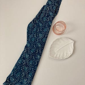 May include: A navy blue scarf with a paisley pattern in light blue and white. A small pink glass and a white leaf-shaped dish are also present on a white surface.
