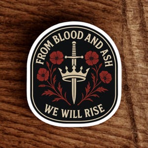 May include: Oval sticker with a black background featuring a sword, crown, and floral elements in cream and red. The text "FROM BLOOD AND ASH" arches across the top, with "WE WILL RISE" at the bottom.