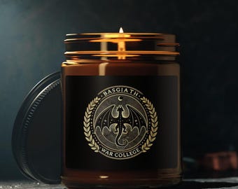 Può includere: Un barattolo di vetro marrone con una candela, con un'etichetta nera decorata con un motivo dorato di un drago con le ali spiegate, una luna crescente e stelle. L'etichetta riporta anche il testo "Basgiath War College".