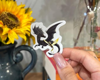 Pode incluir: Um adesivo de um dragão preto e dourado com um dragão dourado menor nas costas. O adesivo está sobre um fundo branco.