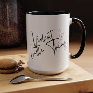 Puede incluir: Taza de cerámica blanca con borde y asa negros. La taza tiene texto negro que dice "Violent Little Thing" con un corazón rosa.