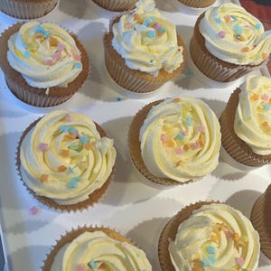May include: A tray of cupcakes, each in a paper case, with swirled yellow buttercream and colourful star sprinkles. The cupcakes are ready to be enjoyed. The image shows a close-up of the baked goods.