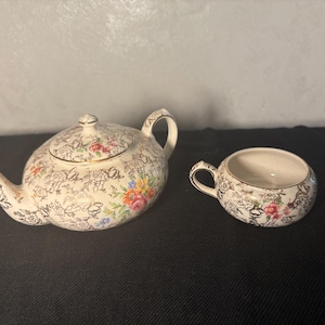 May include: A vintage cream-colored teapot with a matching cup. Both pieces feature a gold filigree design with colorful floral accents. The teapot has a lid and a curved spout. The cup has a small handle.