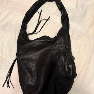 May include: Black leather hobo bag with a knotted handle and a small external pocket. The bag has a soft, slouchy design and a long, knotted strap. The leather appears textured, and the bag has a casual, everyday style.