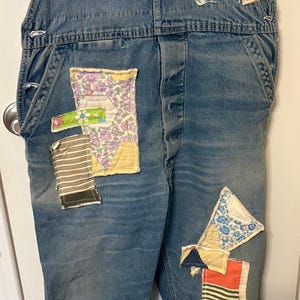 May include: Blue denim overalls with colorful fabric patches. Patches include floral, striped, and solid patterns. A red heart-shaped patch is sewn on the bib. The overalls have metal buttons and adjustable straps.
