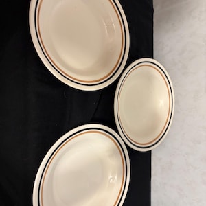 Puede incluir: Tres platos de cerámica blanquecina con un borde negro y marrón. Los platos son de forma ovalada y parecen cuencos o platos. El fondo es una tela negra y una superficie blanca.