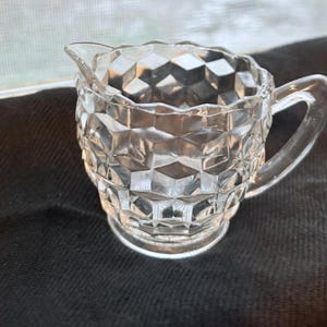 May include: A clear glass creamer with a diamond pattern. The creamer has a handle and a rounded base.