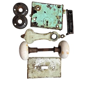 Könnte beinhalten: Sammlung antiker Türbeschlagteile, darunter ein verwittertes türkisblaues Schloss, ein weißer Porzellanknauf und verschiedene Metallplatten. Die Teile weisen Alters- und Gebrauchsspuren auf, mit abblätternder Farbe und Rost.