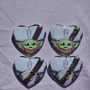 May include: Four heart-shaped pins featuring a green character with large eyes and ears, wearing a brown garment and a dark strap. Two pins have a smooth finish, while the other two display a holographic effect with small heart patterns.