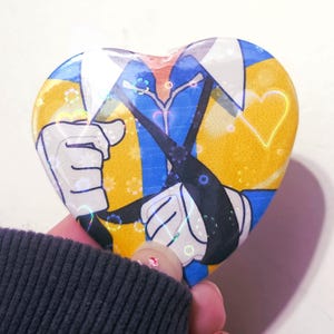 May include: Heart-shaped button with a cartoon illustration. The design features a character holding umbrellas, with a yellow and blue background. The button is held by a person wearing a dark gray sweater.