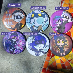 May include: A collection of six round buttons with colorful character designs. The buttons feature various characters with text labels including "Butler N", "Squished N", "Prom Nite", "Uzi A", "Squished Uzi", and "Cyn A". The buttons are arranged on a textured surface.