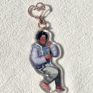 May include: A clear acrylic keychain featuring a seated cartoon figure in a blue and gray spacesuit. The figure has dark curly hair and is wearing sandals. The keychain has a rose gold heart-shaped clasp.