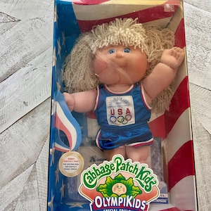 May include: A Cabbage Patch Kids doll wearing a blue and red Olympic uniform with the words "Olympikids 1996" and "USA" on the front. The doll is holding a red, white, and blue ribbon. The doll is in a box with a red, white, and blue American flag design.