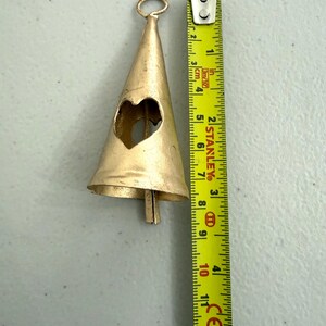 May include: A gold-colored metal bell with a heart-shaped cutout. The bell is approximately 5 inches long, with a loop at the top for hanging. A measuring tape is next to the bell.