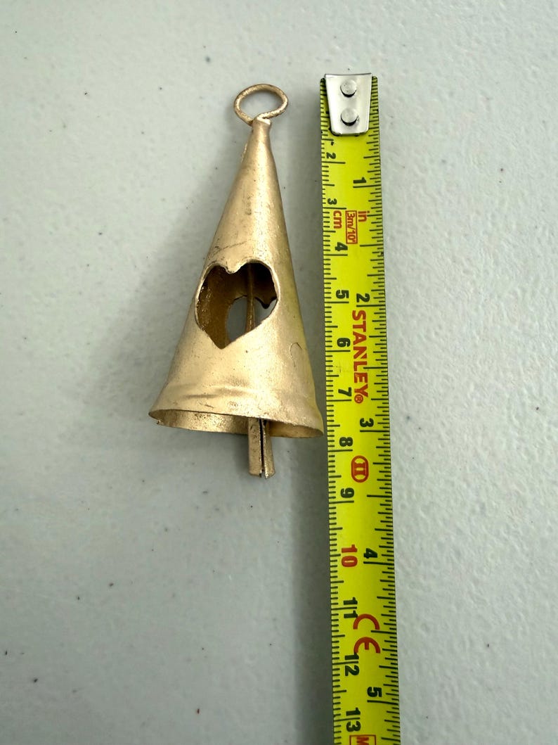May include: A gold-colored metal bell with a heart-shaped cutout. The bell is approximately 5 inches long, with a loop at the top for hanging. A measuring tape is next to the bell.