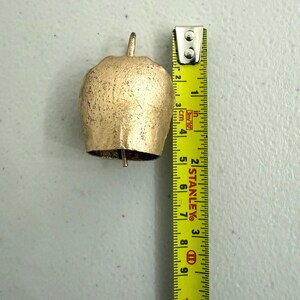May include: A gold-colored bell with a textured surface, approximately 3 inches tall. The bell has a loop at the top and a clapper visible inside. A yellow measuring tape is next to the bell.
