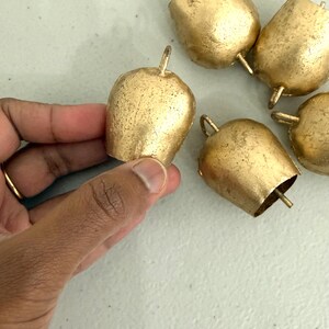 May include: A collection of small, gold-colored bells with a textured surface. One bell is held by a hand, while the others are scattered on a light-colored surface. Each bell has a loop at the top and a flared bottom.