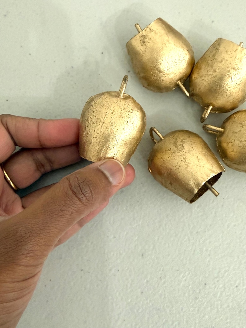 May include: A collection of small, gold-colored bells with a textured surface. One bell is held by a hand, while the others are scattered on a light-colored surface. Each bell has a loop at the top and a flared bottom.