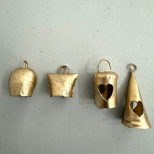 May include: Four antique gold-colored bells of varying shapes. The bells include a rounded bell, a square bell, a bell with a heart-shaped cutout, and a conical bell with a heart-shaped cutout. Each bell has a loop for hanging.