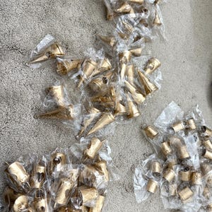 May include: Several small, gold-colored bells in clear plastic bags are scattered across a textured, gray surface. The bells vary in shape, some conical and others cylindrical, suggesting a craft or decorative theme.
