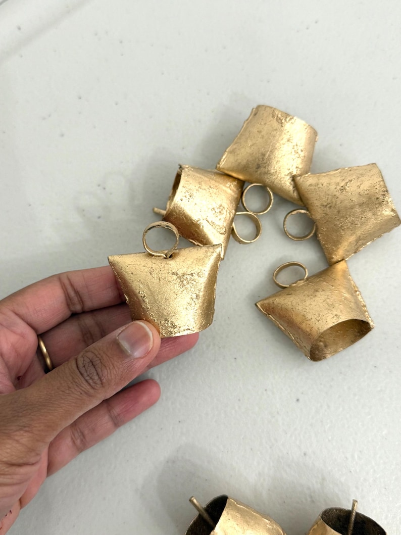 May include: A collection of small, gold-colored metal bells with a textured finish. The bells have a trapezoidal shape with a small loop at the top for hanging. Some bells are scattered, while one is held by a hand.