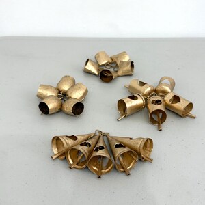 May include: Several clusters of small, antique gold-colored bells. The bells are in various shapes, including rounded and conical, and are arranged on a light gray surface. Each bell has a small clapper inside.