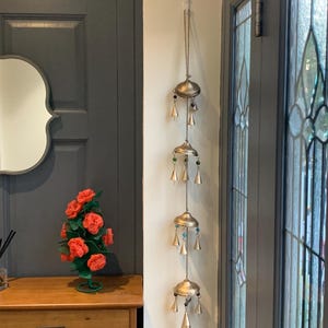 May include: A gold wind chime with multiple tiers of bells hanging from a white ceiling hook. The chime is decorated with small, colorful beads. The chime is hanging in a doorway with a dark gray door and a white wall.