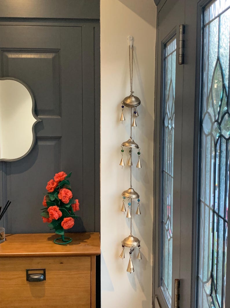 May include: A gold wind chime with multiple tiers of bells hanging from a white ceiling hook. The chime is decorated with small, colorful beads. The chime is hanging in a doorway with a dark gray door and a white wall.