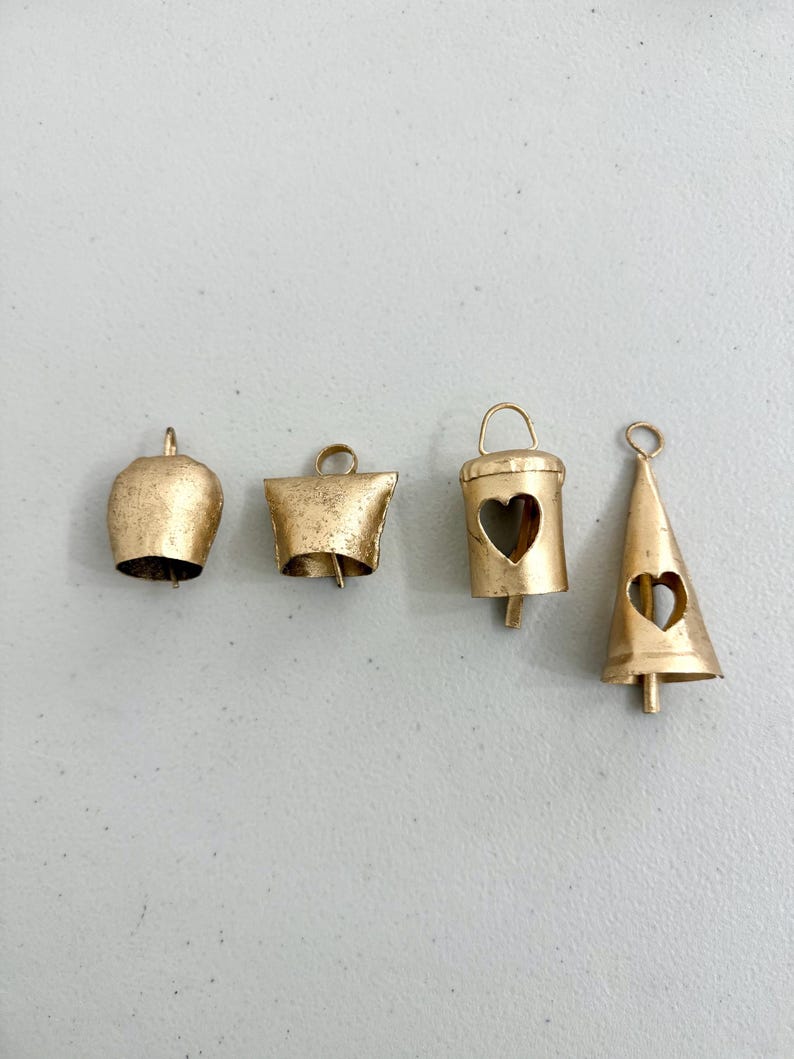 May include: Four gold-colored metal bells of varying shapes. The bells include a rounded bell, a square bell, a cylindrical bell with a heart cutout, and a conical bell with a heart cutout. Each bell has a loop for hanging.