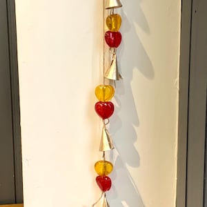 Puede incluir: Una cadena de cuentas de corazón rojas y amarillas con campanas plateadas que cuelgan de una cuerda de yute.