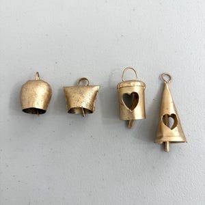 May include: Four small, antique gold-colored bells in various shapes. The bells include a classic bell shape, a flared bell, a bucket-shaped bell with a heart cutout, and a cone-shaped bell with a heart cutout. Each bell has a loop for hanging.