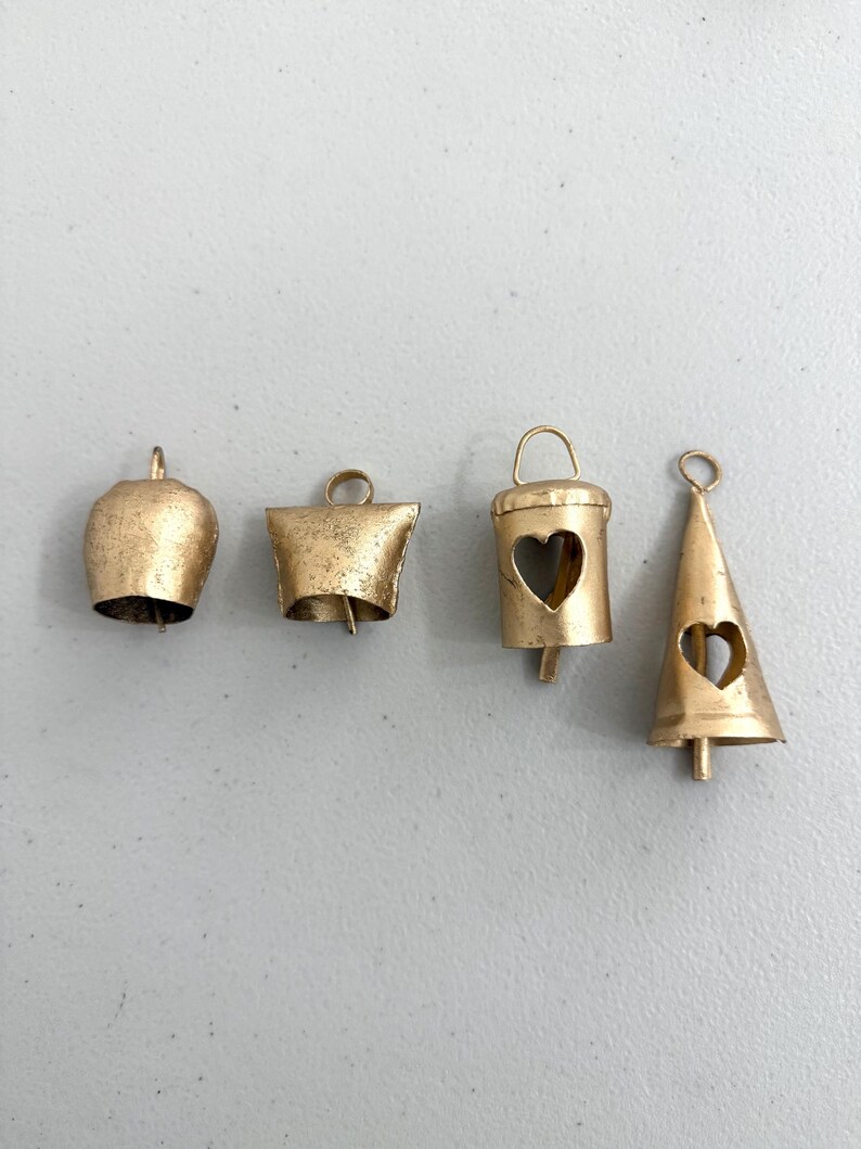 May include: Four small, antique gold-colored bells in various shapes. The bells include a classic bell shape, a flared bell, a bucket-shaped bell with a heart cutout, and a cone-shaped bell with a heart cutout. Each bell has a loop for hanging.