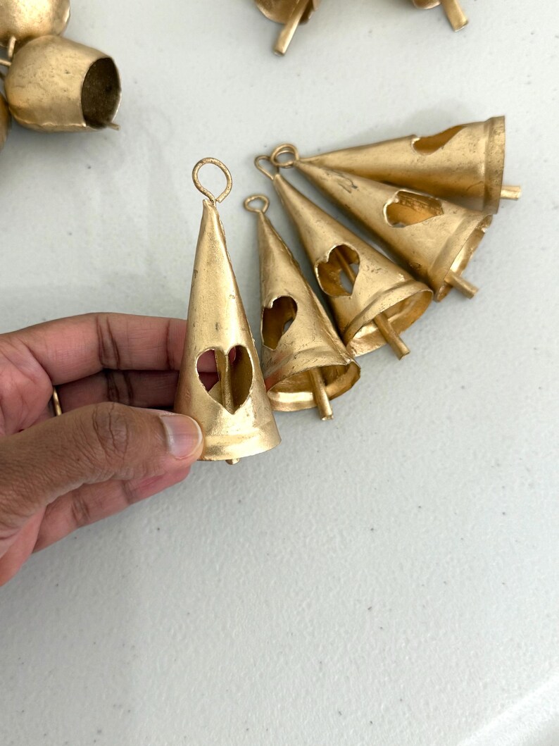 May include: A collection of gold-colored metal bells. The bells are cone-shaped with a heart-shaped cutout and a loop at the top for hanging. Some bells are in focus, while others are blurred in the background.