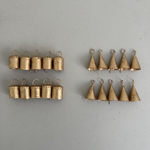 Può includere: Due serie di piccole campane color oro. Una serie presenta campane cilindriche con anelli, l'altra campane a forma di cono. Le campane sono disposte su una superficie grigio chiaro, ideali per l'artigianato o la decorazione.