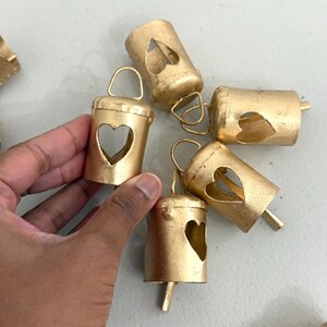 May include: Gold-colored metal bells with heart-shaped cutouts. Each bell has a small loop at the top and a clapper. The bells are arranged on a light gray surface.