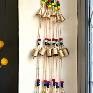 Puede incluir: Adorno colgante decorativo con múltiples campanas pequeñas de color dorado suspendidas de hilo de yute. El hilo está adornado con cuentas de colores rojo, verde, amarillo y negro. Las campanas están dispuestas en cascada.