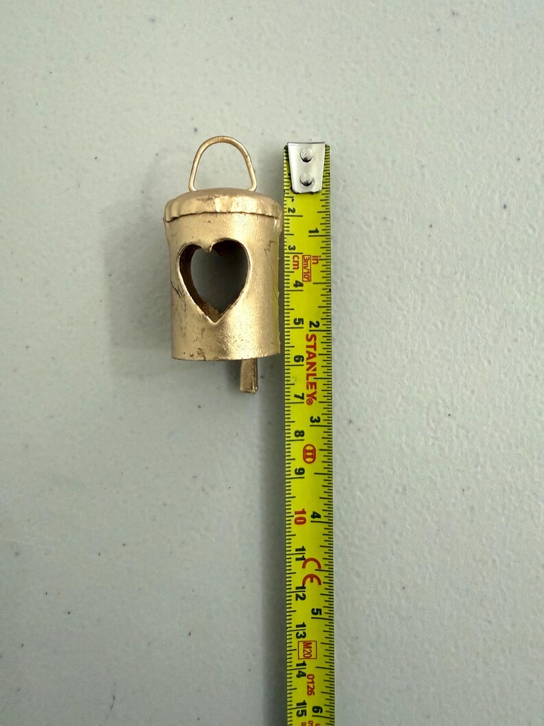May include: A small, gold-colored bell with a heart-shaped cutout. The bell has a loop at the top and a clapper. The bell is approximately 3 inches tall, as measured by a yellow tape measure.