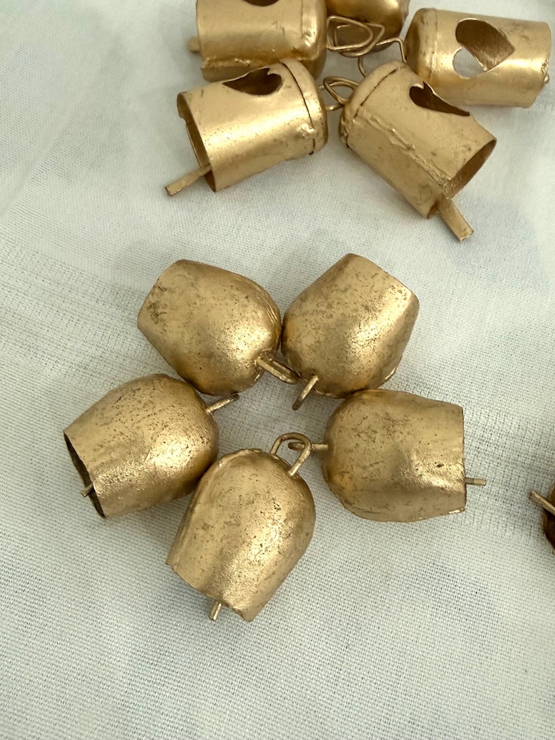 May include: A collection of small, gold-colored metal bells. Some bells have a heart-shaped cutout. The bells are arranged on a white, textured surface. The bells have a rustic, vintage appearance.