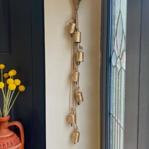 May include: A gold-coloured metal bell garland with a jute rope hanging from a hook on a white wall. The garland has eight bells and is hanging next to a dark wooden door.