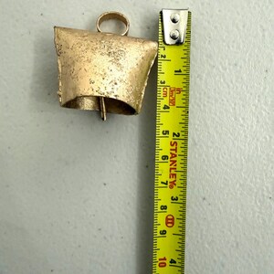 May include: A rustic, gold-colored bell with a loop for hanging. The bell is approximately 4 inches tall. A yellow measuring tape is placed next to the bell, showing the measurement in inches.