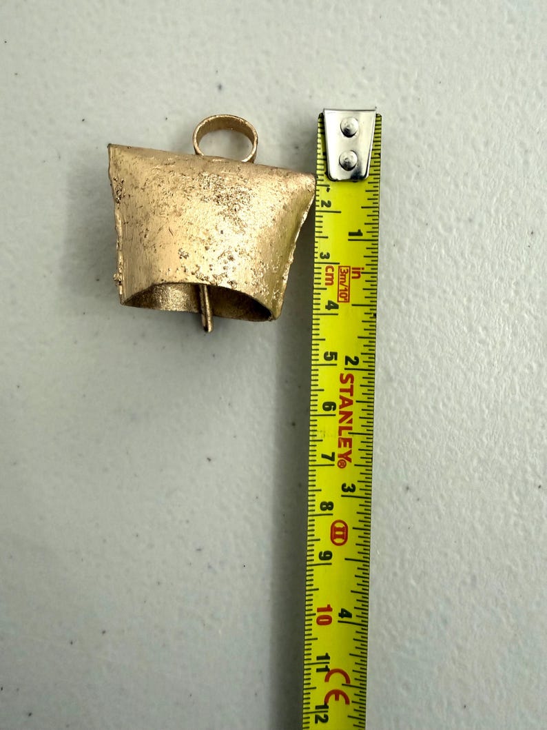 May include: A rustic, gold-colored bell with a loop for hanging. The bell is approximately 4 inches tall. A yellow measuring tape is placed next to the bell, showing the measurement in inches.