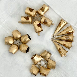 May include: An assortment of antique gold-colored bells in various shapes, including heart-cutout cylinders, rounded bells, and cone-shaped bells. The bells are arranged on a white, textured surface, suggesting a craft or decorative theme.