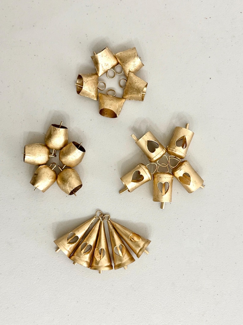 May include: An assortment of small, gold-colored bells in various arrangements. Some bells have heart-shaped cutouts. The bells are clustered in groups, with some arranged in a circular pattern. The overall aesthetic is rustic and decorative.