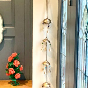 May include: A silver wind chime with four tiers of bells and colorful beads hanging from a white wall. The chime is made of metal and has a rustic look.