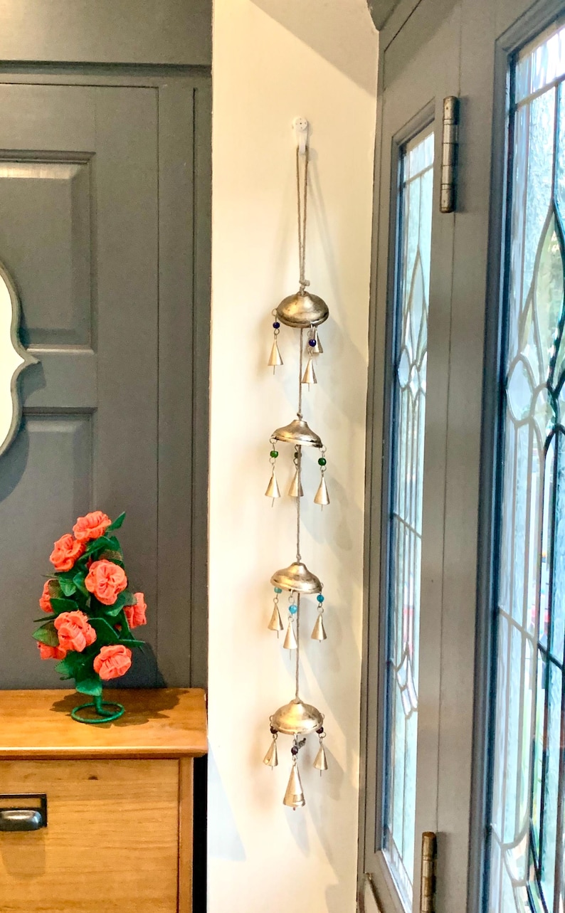 May include: A silver wind chime with four tiers of bells and colorful beads hanging from a white wall. The chime is made of metal and has a rustic look.