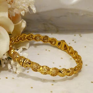 Könnte beinhalten: Ein goldenes Armreif-Armband mit einem zarten, filigranen Design. Das Armband besteht aus einer Reihe kleiner, miteinander verbundener Kreise.