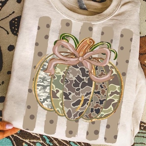 Op de afbeelding: Een beige sweater met ronde hals en een centrale afbeelding van een pompoen met een camouflagepatroon en een lichtbruine strik. De pompoen is geplaatst over een achtergrond van verticale strepen met bruine stippen op een beige stof.