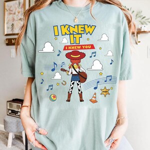 May include: A light blue t-shirt featuring a graphic of a cowgirl character playing a guitar. The text reads I KNEW IT and I KNEW YOU. The design includes musical notes, clouds, stars, a ball, a traffic cone, and a sheriff badge, all in a cartoon style.